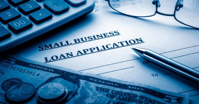 A small business loan application sitting on a desk. Nearby is a calculator, a pen, a pair of glasses, and some cash.