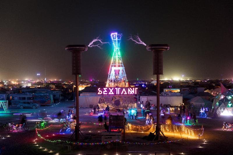 A nighttime capture of dual resonant transformer coils (Tesla coils) flanking an illuminated central tower at Burning Man’s Sextant Camp as shown on Smith’s LinkedIn profile.