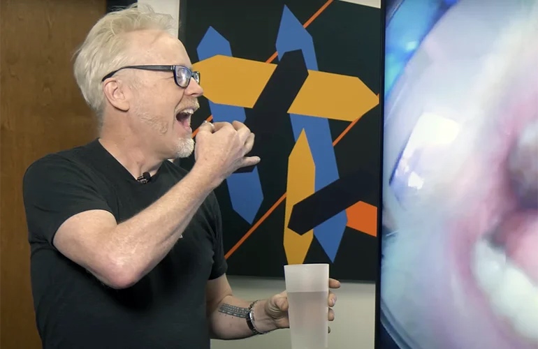 Adam Savage holding a PillBot from Endiatx up to his open mouth. In his other hand, he holds a glass of water. 